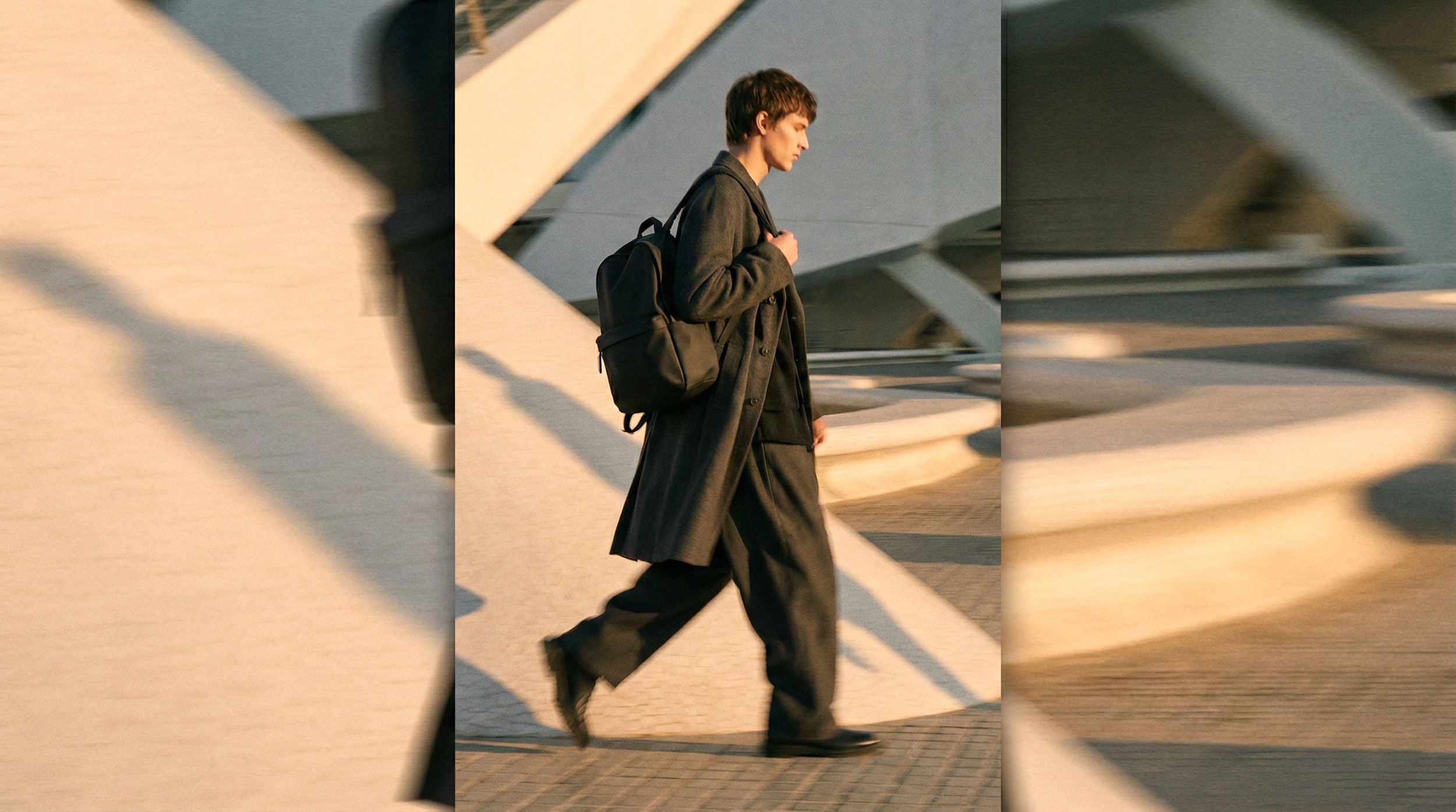 Fotografía de lifestyle urbano en movimiento con estética street style. Un hombre viste abrigo gris largo y pantalones anchos mientras camina llevando una mochila negra minimalista NVMA en la Ciudad de las Artes y las Ciencias (Valencia). Iluminación de atardecer con sombras arquitectónicas marcadas.