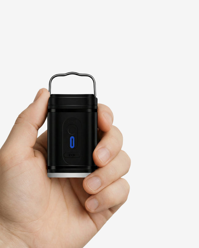 Mano sosteniendo el mini compresor eléctrico portátil V-Flow negro de NVMA. Vista de cerca que destaca su diseño ultra compacto, ergonómico y ligero, con el indicador LED azul de encendido activado.
